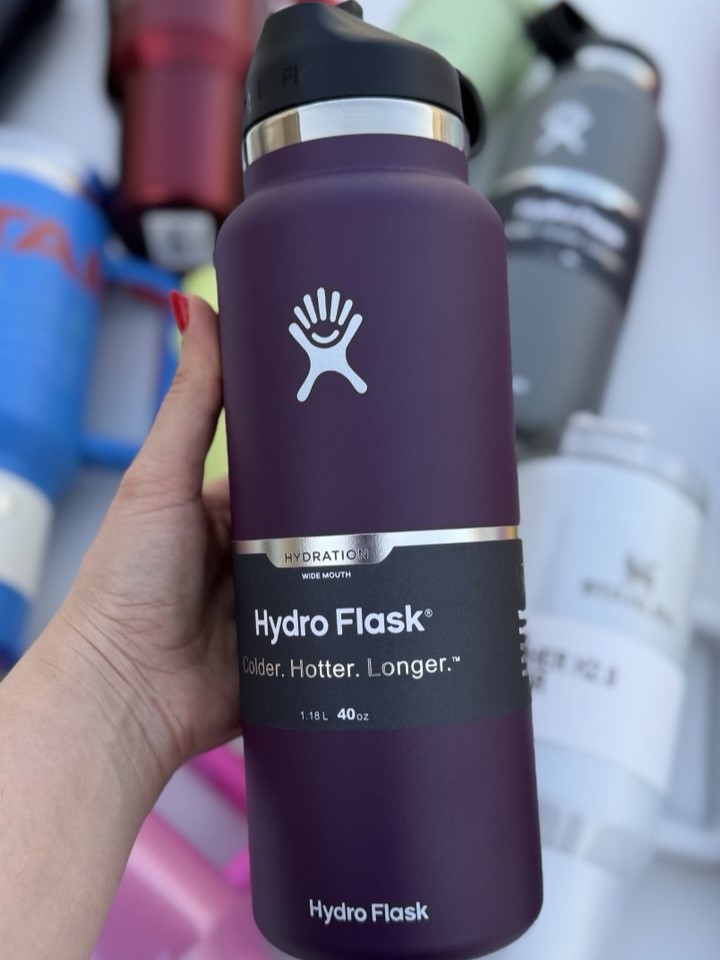 بطری بنفش تیره Hydro Flask با در مشکی و نشان سفید، در دست و مقابل بطری‌های دیگر